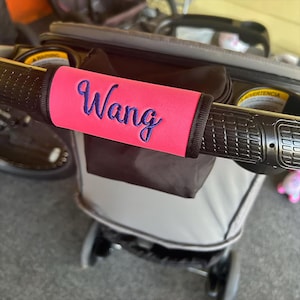 May include: A pink stroller handle cover with the name "Wang" embroidered in blue script. The cover is wrapped around a black stroller handle, with a black trim. The stroller has a black and gray color scheme.