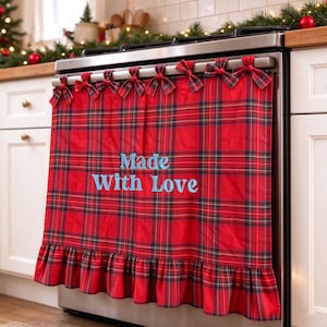 May include: A red tartan stove cover featuring a ruffled bottom edge and seven small fabric bows along the top. The text Made With Love is embroidered in light blue thread across the centre of the fabric, which hangs over the front of a stove.