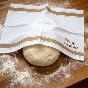 May include: A white kitchen towel with two horizontal tan stripes and the embroidered brown text Life of a Dough Girl, draped over a round ball of raw bread dough. The towel features a clean, rectangular design with hemmed edges.