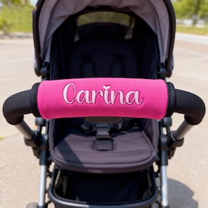 May include: A hot pink stroller handlebar cover with the name "Carina" embroidered in white script. The cover is wrapped around a black handlebar, with a black stroller in the background.