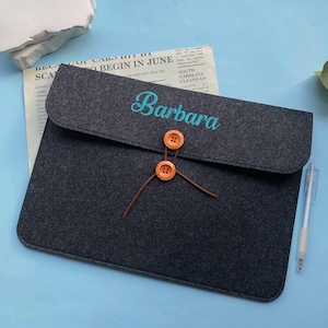 Puede incluir: Una funda para portátil de fieltro gris oscuro con el nombre "Barbara" bordado en turquesa. La funda tiene un cierre de solapa asegurado por dos botones de madera naranja y una cuerda marrón. Un bolígrafo descansa sobre la superficie azul.