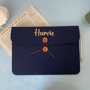 May include: A navy blue felt document case with the name "Harvie" embroidered in gold. The case has a flap closure secured by two brown buttons and a string. A newspaper is visible in the background.