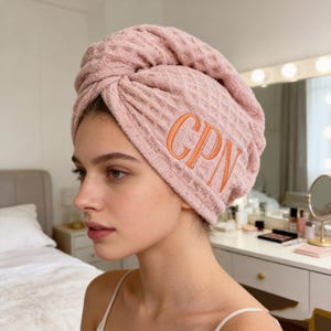 May include: A light pink waffle-weave hair towel wrapped around the head, embroidered with the letters "CPN" in orange. The towel is made of a soft, absorbent material, designed to dry hair quickly and comfortably. The towel is a stylish and practical accessory.
