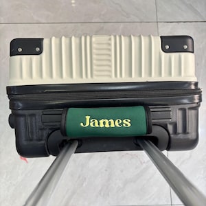Op de afbeelding: Een zwarte en crèmekleurige rolkoffer met een groene handgreep. De naam "James" is in goud op de handgreep geborduurd. De koffer heeft een geribbeld ontwerp aan de bovenkant en zwarte hoekbeschermers.
