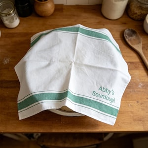 May include: A white kitchen towel with two horizontal green stripes draped over a bowl. The fabric features the embroidered text "Abby's Sourdough" in a matching green font. The towel is made of a woven material and covers the contents of the bowl.