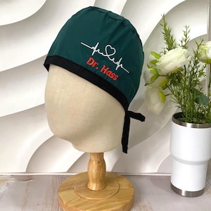 May include: A dark green scrub cap featuring a black trim and tie-back closure. The cap is embroidered with a white EKG heartbeat line that forms a heart shape, positioned above the red embroidered text that reads Dr. Hass.