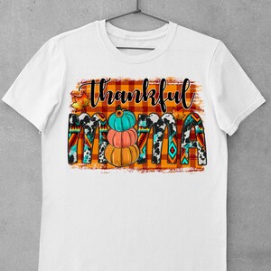 K&ouml;nnte beinhalten: Wei&szlig;es T-Shirt mit dem Text "Thankful Mama" in schwarz-wei&szlig;en Buchstaben auf kariertem Hintergrund. Der Text ist mit Herbstbl&auml;ttern und K&uuml;rbissen verziert.