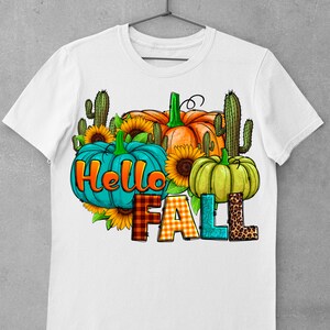 May include: White t-shirt with a graphic design featuring pumpkins, sunflowers, and cacti. The text "Hello Fall" is written in a colorful, patterned font.