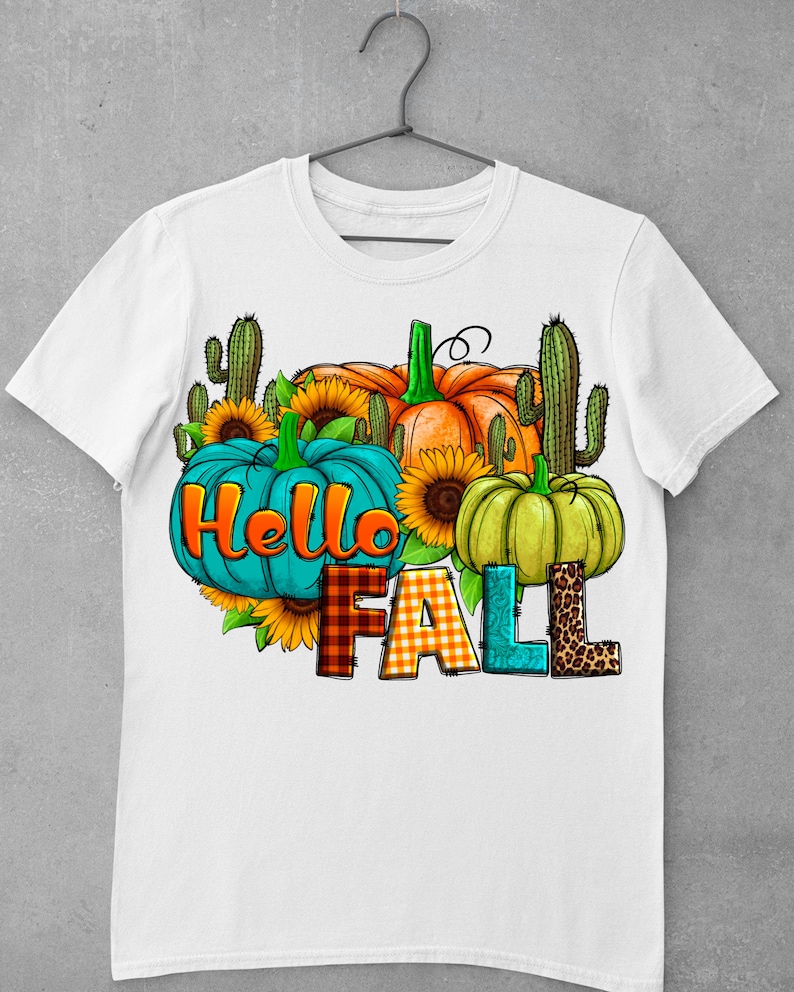 May include: White t-shirt with a graphic design featuring pumpkins, sunflowers, and cacti. The text "Hello Fall" is written in a colorful, patterned font.