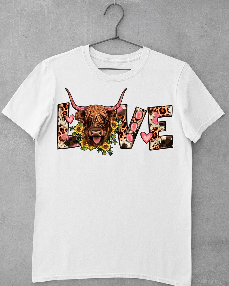 May include: White t-shirt featuring a graphic of a Highland cow with a floral wreath around its head. The word "LOVE" is spelled out in large letters with a leopard print and pink hearts.