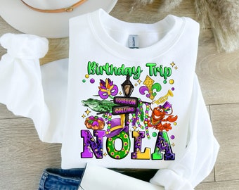 May include: White sweatshirt with a green, purple, and gold graphic design that says "Birthday Trip" and "NOLA". The graphic includes a Mardi Gras mask, a street lamp, a fleur-de-lis, a trumpet, a crawfish, and a sign that says "Bourbon Orleans".