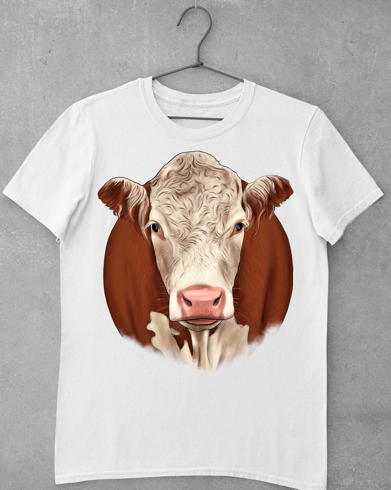 May include: White t-shirt featuring a detailed illustration of a Hereford cow's face. The cow has a white face with brown markings around the eyes and ears, and a pink nose. The artwork is set against a brown background.