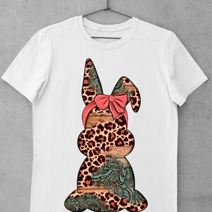 May include: White t-shirt with a graphic of a brown and orange leopard print bunny wearing a pink bow. The bunny is sitting on a wooden background with a turquoise floral pattern.