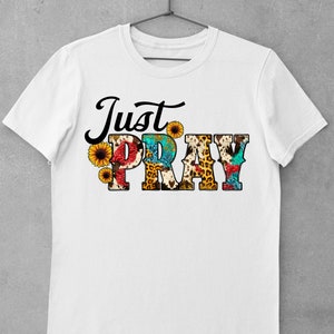 May include: White t-shirt with the text "Just Pray" in black lettering. The letters are decorated with a variety of colorful patterns, including leopard print, sunflowers, and paisley.