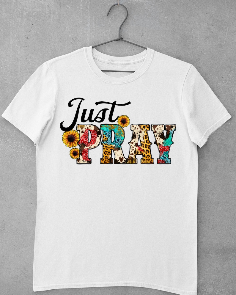 May include: White t-shirt with the text "Just Pray" in black lettering. The letters are decorated with a variety of colorful patterns, including leopard print, sunflowers, and paisley.