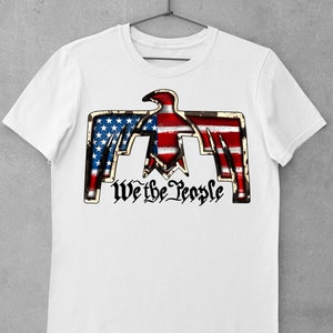 May include: White t-shirt with a graphic of a stylized eagle with the American flag design. The eagle is holding the words "We the People" in a stylized font.