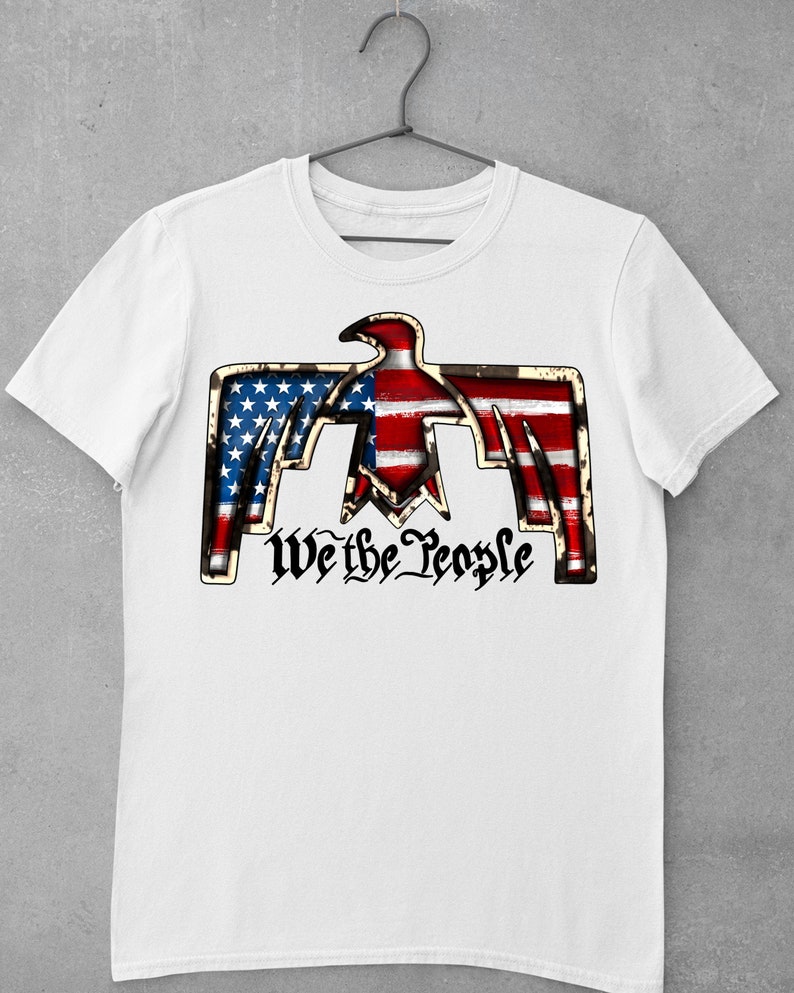 May include: White t-shirt with a graphic of a stylized eagle with the American flag design. The eagle is holding the words "We the People" in a stylized font.