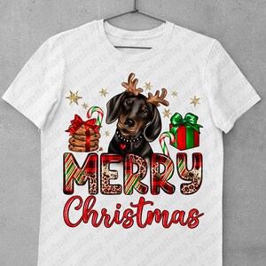 May include: A white t-shirt with a black and brown dachshund wearing a red and green plaid Santa hat with antlers. The dog is holding a candy cane and a gift box. The text "Merry Christmas" is written in red and green plaid with leopard print accents.