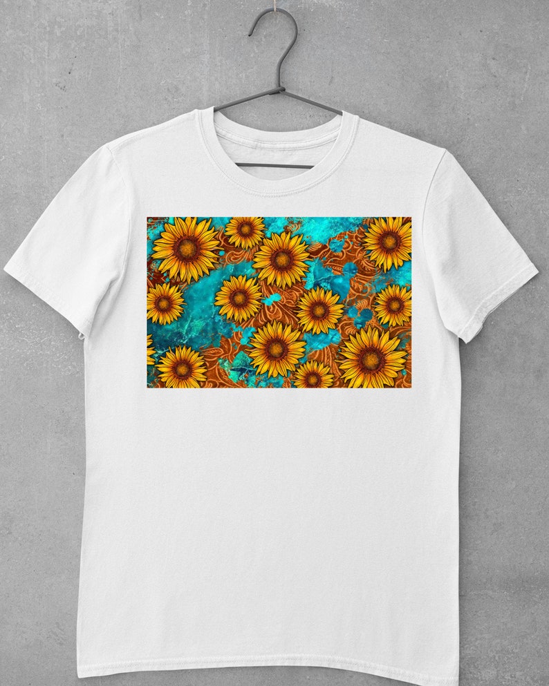May include: White t-shirt featuring a vibrant sunflower design. The graphic showcases yellow sunflowers with brown centers against a teal and brown patterned background. The shirt is displayed on a hanger.