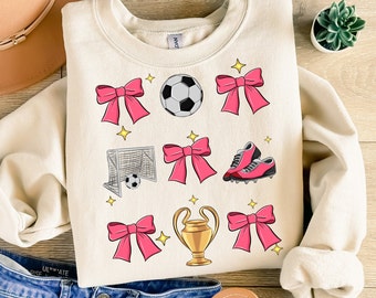 Könnte beinhalten: Ein cremefarbener Pullover mit einem pink-weißen Fußball-Motiv. Das Design beinhaltet einen Fußball, ein Tor, Fußballschuhe und Schleifen.