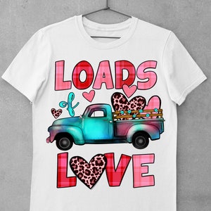 May include: White t-shirt featuring a teal and pink truck carrying hearts. The text "LOADS of LOVE" is displayed in pink and red, with heart accents. The truck bed has a leopard print heart. A Valentine's Day themed design.