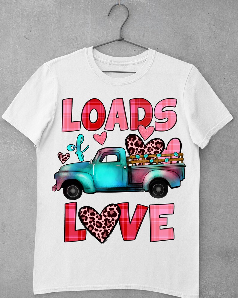 May include: White t-shirt featuring a teal and pink truck carrying hearts. The text "LOADS of LOVE" is displayed in pink and red, with heart accents. The truck bed has a leopard print heart. A Valentine's Day themed design.