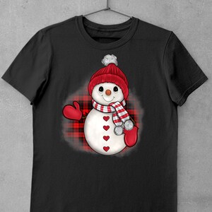 May include: Black t-shirt with a snowman design. The snowman is wearing a red knitted hat with a white bobble, a striped scarf, and red mittens. The snowman's body has red heart accents. The design is set against a red and black plaid background.