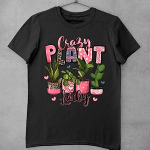 Pu&ograve; includere: Maglietta nera con il testo "Crazy Plant Lady" in lettere rosa. Il testo &egrave; circondato da cuori rosa e una fila di piante in vaso con vasi a motivi rosa e bianchi.