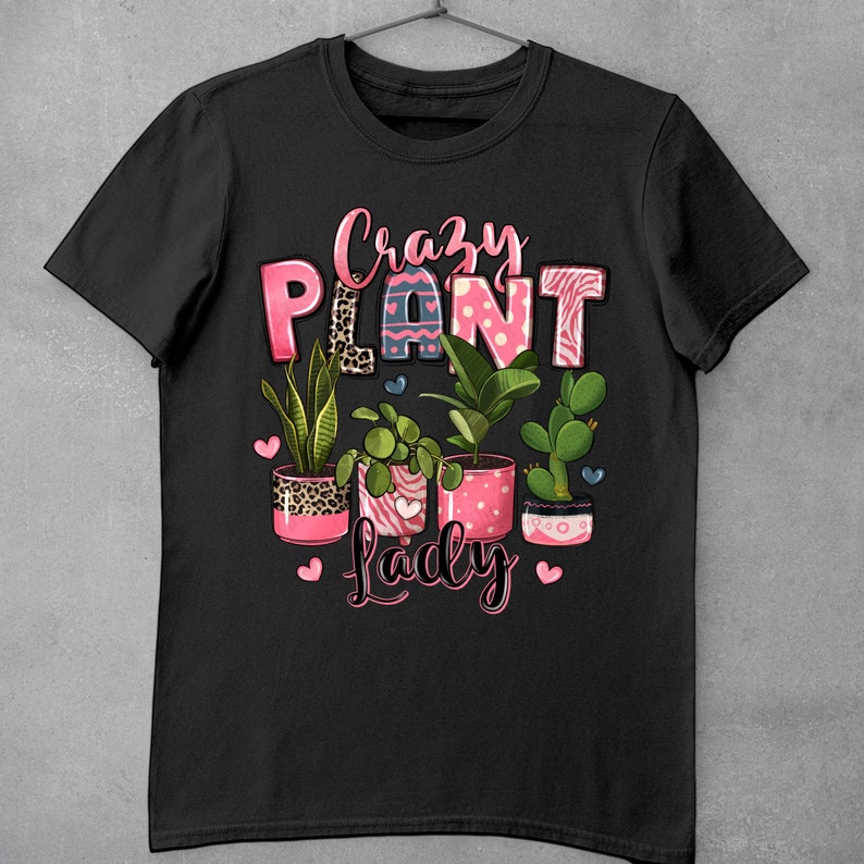 Pu&ograve; includere: Maglietta nera con il testo "Crazy Plant Lady" in lettere rosa. Il testo &egrave; circondato da cuori rosa e una fila di piante in vaso con vasi a motivi rosa e bianchi.