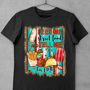 Puede incluir: Camiseta negra con un dise&ntilde;o gr&aacute;fico que presenta una variedad de alimentos callejeros, incluyendo un ma&iacute;z en la mazorca, churros, un taco y una paleta de hielo. El texto "Feed me street food and tell me I'm pretty" est&aacute; impreso en la camiseta.