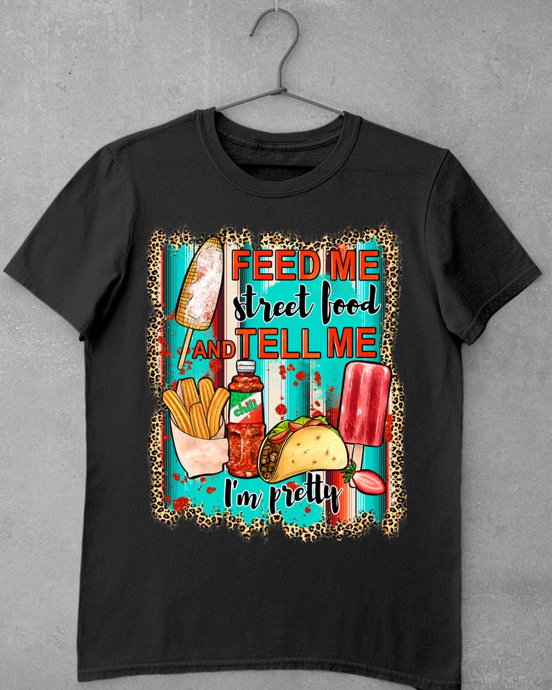 Puede incluir: Camiseta negra con un dise&ntilde;o gr&aacute;fico que presenta una variedad de alimentos callejeros, incluyendo un ma&iacute;z en la mazorca, churros, un taco y una paleta de hielo. El texto "Feed me street food and tell me I'm pretty" est&aacute; impreso en la camiseta.