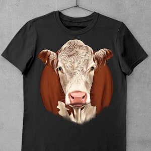 May include: Black t-shirt featuring a detailed illustration of a cow's face. The cow has a white and light brown face with a pink nose and brown ears. The cow's head is set against a brown circular background.