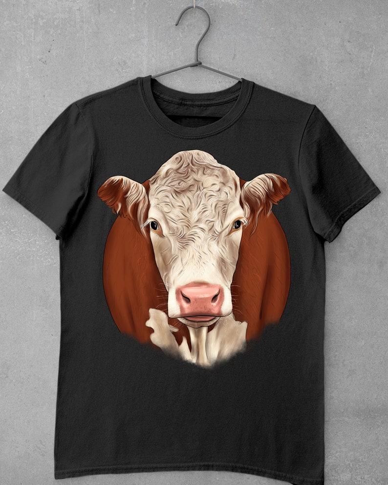 May include: Black t-shirt featuring a detailed illustration of a cow's face. The cow has a white and light brown face with a pink nose and brown ears. The cow's head is set against a brown circular background.