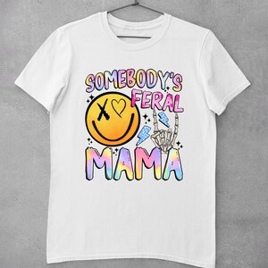 Puede incluir: Camiseta blanca con un dise&ntilde;o gr&aacute;fico colorido que dice "Somebody's Feral Mama". El gr&aacute;fico incluye una cara sonriente con una X para un ojo y un coraz&oacute;n para el otro ojo, una mano de esqueleto con un rayo y un texto "Mama" en colores arco&iacute;ris.