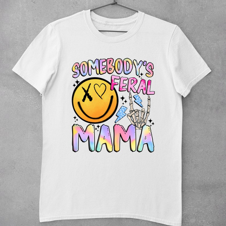 Puede incluir: Camiseta blanca con un dise&ntilde;o gr&aacute;fico colorido que dice "Somebody's Feral Mama". El gr&aacute;fico incluye una cara sonriente con una X para un ojo y un coraz&oacute;n para el otro ojo, una mano de esqueleto con un rayo y un texto "Mama" en colores arco&iacute;ris.