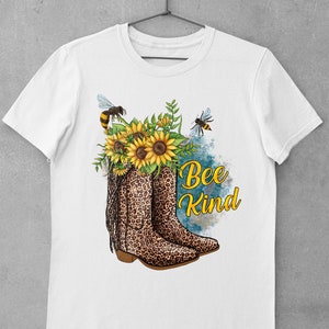May include: White t-shirt with a graphic of a pair of brown and black leopard print cowboy boots with yellow sunflowers and bees. The text "Bee Kind" is written in black lettering.