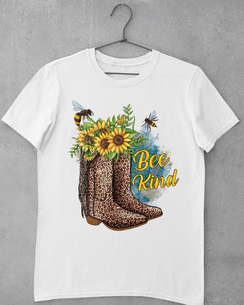 May include: White t-shirt with a graphic of a pair of brown and black leopard print cowboy boots with yellow sunflowers and bees. The text "Bee Kind" is written in black lettering.