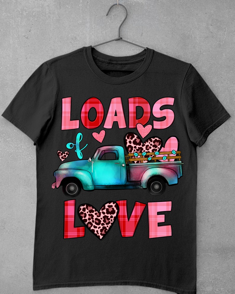 May include: Black t-shirt featuring a teal and pink vintage truck carrying leopard print hearts. The words "LOADS of LOVE" are in pink and red plaid lettering. Small pink hearts are scattered around the truck design.
