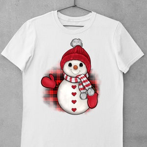 May include: White t-shirt with a cartoon snowman design. The snowman has a red knitted hat with a white bobble, a striped scarf, and red mittens. The snowman's body has red heart-shaped buttons. A red and black plaid pattern is behind the snowman.