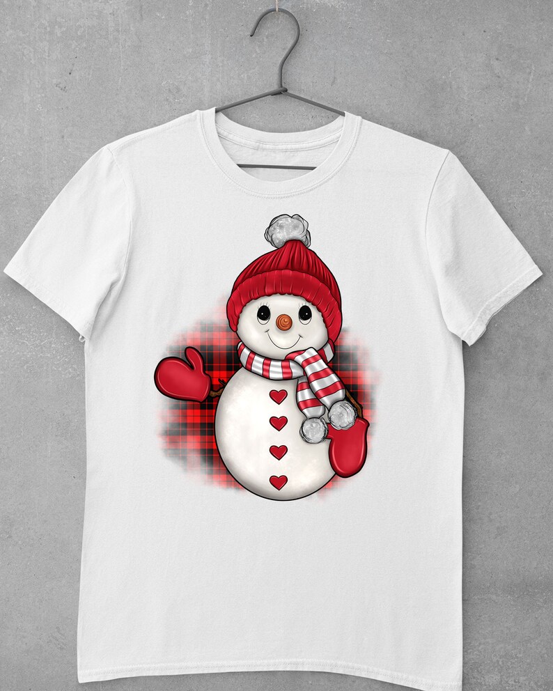 May include: White t-shirt with a cartoon snowman design. The snowman has a red knitted hat with a white bobble, a striped scarf, and red mittens. The snowman's body has red heart-shaped buttons. A red and black plaid pattern is behind the snowman.