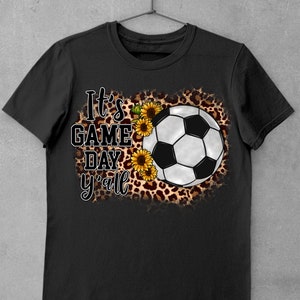 Puede incluir: Camiseta negra con un bal&oacute;n de f&uacute;tbol blanco, girasoles y estampado de leopardo. El texto "It's Game Day y'all" est&aacute; impreso en la camiseta.