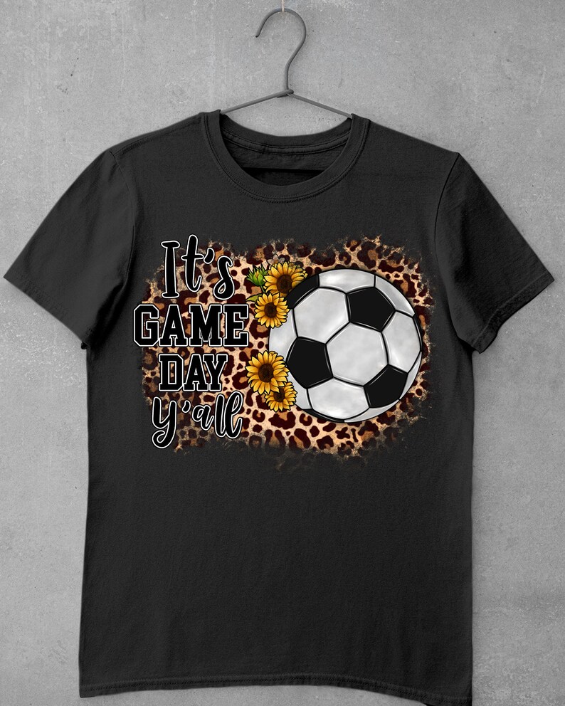 Puede incluir: Camiseta negra con un bal&oacute;n de f&uacute;tbol blanco, girasoles y estampado de leopardo. El texto "It's Game Day y'all" est&aacute; impreso en la camiseta.