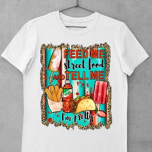 Puede incluir: Camiseta blanca con un dise&ntilde;o gr&aacute;fico que presenta una variedad de alimentos callejeros, incluyendo churros, tacos, paletas de hielo y una botella de salsa picante. El texto en la camiseta dice "Feed me street food and tell me I'm pretty."