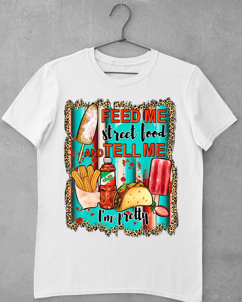 Puede incluir: Camiseta blanca con un dise&ntilde;o gr&aacute;fico que presenta una variedad de alimentos callejeros, incluyendo churros, tacos, paletas de hielo y una botella de salsa picante. El texto en la camiseta dice "Feed me street food and tell me I'm pretty."