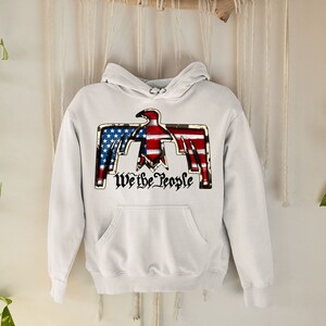 May include: White hooded sweatshirt with a graphic of an eagle with the American flag design and the text "We the People".