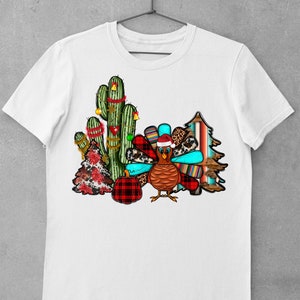 May include: A white t-shirt featuring a festive design with a turkey wearing a Santa hat, a cactus decorated with Christmas lights, a plaid pumpkin, and a Christmas tree with a red and white plaid pattern.