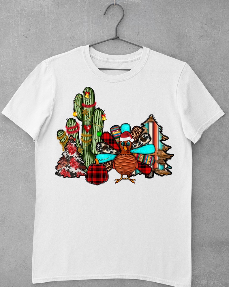 May include: A white t-shirt featuring a festive design with a turkey wearing a Santa hat, a cactus decorated with Christmas lights, a plaid pumpkin, and a Christmas tree with a red and white plaid pattern.