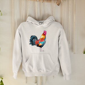 May include: A white hoodie with a colorful rooster illustration on the front. The rooster has a red comb, yellow and orange feathers, and blue and black tail feathers.