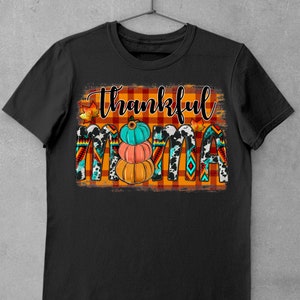 K&ouml;nnte beinhalten: Schwarzes T-Shirt mit einem grafischen Design des Wortes "thankful" in wei&szlig;er Schrift auf einem rot-orangefarbenen karierten Hintergrund. Das Wort "mama" ist in wei&szlig;er Schrift auf einem schwarz-wei&szlig;en Kuhmuster-Hintergrund. Es gibt drei K&uuml;rbisse im Design, einen orangefarbenen, einen pfirsichfarbenen und einen t&uuml;rkisfarbenen.