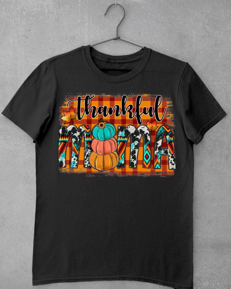 K&ouml;nnte beinhalten: Schwarzes T-Shirt mit einem grafischen Design des Wortes "thankful" in wei&szlig;er Schrift auf einem rot-orangefarbenen karierten Hintergrund. Das Wort "mama" ist in wei&szlig;er Schrift auf einem schwarz-wei&szlig;en Kuhmuster-Hintergrund. Es gibt drei K&uuml;rbisse im Design, einen orangefarbenen, einen pfirsichfarbenen und einen t&uuml;rkisfarbenen.